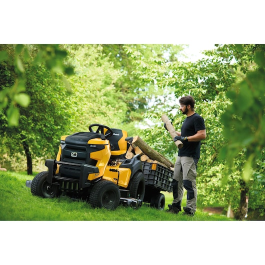CubCadet 14AFA5TQ603 lawn tractor with attached trailer carrying logs, outdoor garden setting.