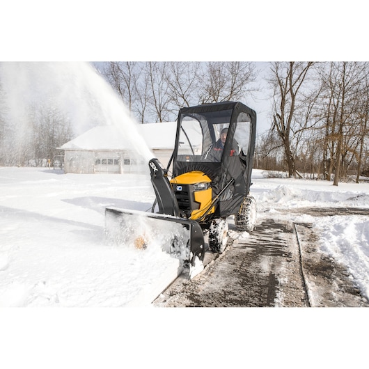 CubCadet 19A40024100 snow blower attachment in use on yellow riding mower with cab, clearing snow from driveway.