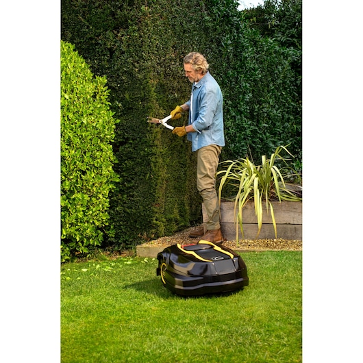 CubCadet 22AKHEFA603 robotic lawn mower on grass in front of person trimming hedge, outdoor garden scene.