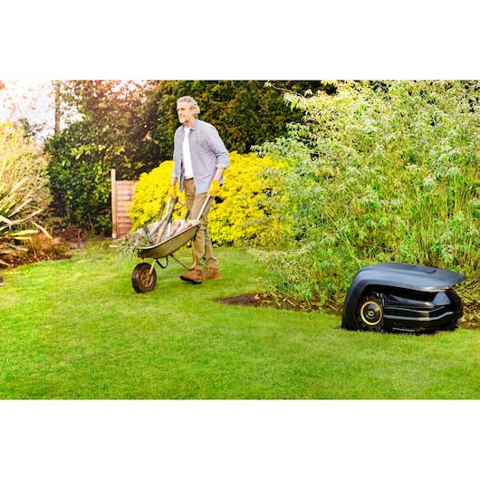 CubCadet 22AKHEFA603 robotic lawn mower in garden near bushes, person with wheelbarrow on grass.