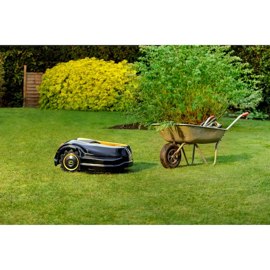 Photo of CubCadet 22AKHEFA603 robotic lawn mower on grass next to a wheelbarrow with garden tools.