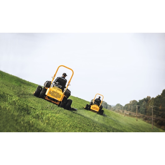 CubCadet 53ALHMUY603 stand-on lawn mowers mowing grass on a sloped field, rear view outdoors.